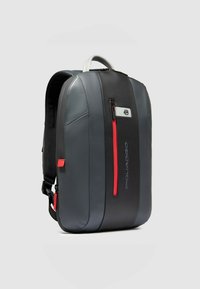 Sac à dos gris et noir en cuir lisse, avec des accents rouges et une poche latérale zippée. Design structuré avec des bretelles ajustables.