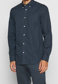 Camicia abbottonata in tessuto blu navy con un piccolo motivo ripetuto di cerchi blu. Design con colletto, bottoni bianchi e maniche lunghe.