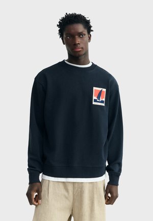 Junger Mann mit kurzen lockigen Haaren, der einen marineblauen Pullover mit Segelboot-Motiv auf der Brust und beige Hosen vor einem einfarbigen Hintergrund trägt.