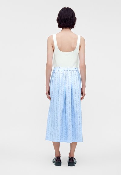 Femme aux cheveux courts et foncés, portant un haut blanc sans manches et un pantalon large bleu clair avec un petit motif à pois, debout de dos.