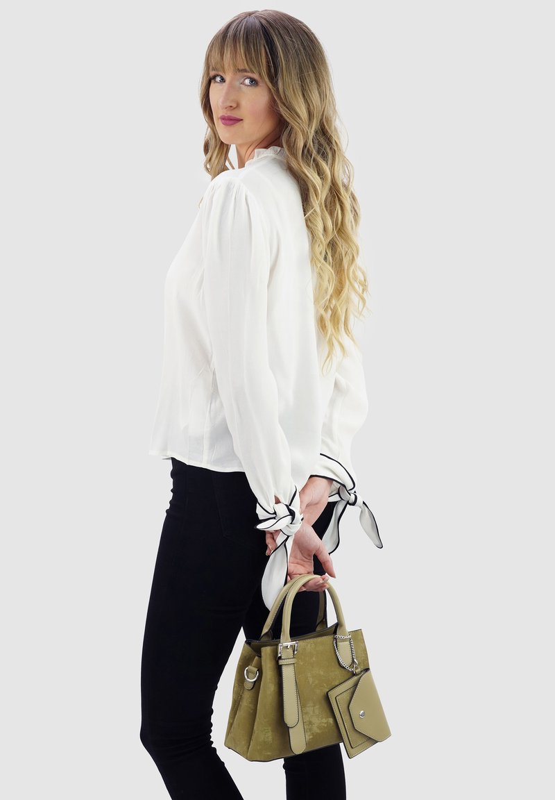 Khaki-Handtasche mit zwei oberen Griffen, Wildledertextur, quadratische Form, ergänzt mit einer abnehmbaren Tasche und silbernem Zubehör. Weiße Bluse und schwarze Hose.