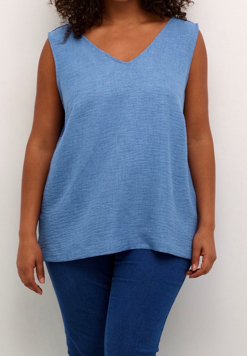 Femme portant un haut bleu clair sans manches à col en V et un pantalon bleu foncé, debout devant un fond clair uni.