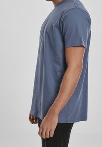 Muž v jednoduchém modrém tričku s krátkým rukávem a černých kalhotách, zobrazený z boku na jednolitém šedém pozadí.