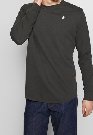 Personne portant un t-shirt gris foncé à manches longues avec un petit logo sur la poitrine et un jean bleu, main touchant le cou sur un fond uni.