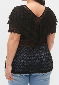 Svart spetsblus med vågiga ärmar och V-bakdesign, med en genomskinlig textur och lager av tyg, ihop med blå denimjeans.