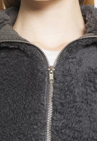 Chaqueta gris de forro polar con cremallera y superficie texturizada y peluda. Presenta una cremallera metálica y un cuello en contraste.