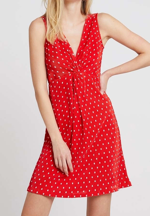 Rotes ärmelloses Kleid mit einem V-Ausschnitt, gerafftem Knoten-Detail an der Vorderseite und weißen Punkten auf einem strukturierten Stoff. Ausgestellte A-Linie.
