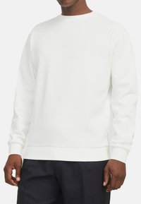 Mannequin in een effen witte lange mouwen crewneck sweatshirt en zwarte broek, staand tegen een witte achtergrond.