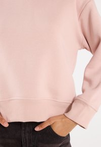 Sweat-shirt rose clair avec une texture douce, des poignets et un ourlet côtelés. Présente une coupe décontractée, avec les mains placées dans des poches en denim noir.