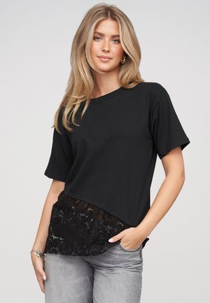 Mujer rubia con una camiseta negra suelta de manga corta con detalle de encaje en el dobladillo y jeans gris claro, de pie frente a un fondo liso.
