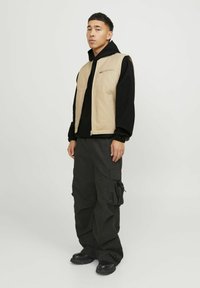 Beige ærmeløs vest over en sort hoodie, båret med mørke cargo-bukser. Struktureret stof med lynlås i siden og lommer. Sorte sko.