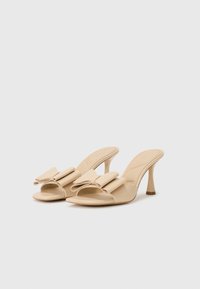 Beige høyhælte mules med en bred sløyfe på fronten, åpen tå-design og en teksturert finish på materialet.