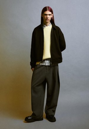 Fekete cipzáras dzseki sárga kötött pulóver és kockás ing felett. Szürke csíkos nadrág és fekete platform cipők fűzővel.