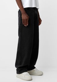 Vaqueros de mezclilla negros holgados con una cintura alta, con detalles de costura mínimos, combinados con zapatillas blancas. Textura suave.