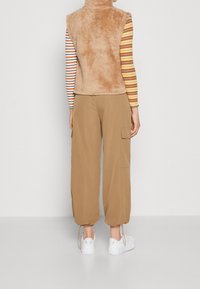 Flauschige beige Weste mit hohem Kragen über einem gestreiften Langarmshirt, kombiniert mit lockeren beige Cargo-Hosen und weißen Turnschuhen.