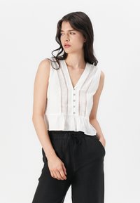 Hvid ærmeløs bluse med V-hals, lodrette striber, blonde detaljer og knaplukning. Peplum kant, båret med sorte snoretræksbukser.