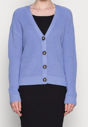 Femme portant un cardigan tricoté violet clair avec quatre boutons sur une robe noire côtelée, debout devant un fond uni.