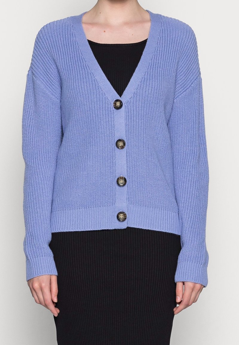 Femme portant un cardigan tricoté violet clair avec quatre boutons sur une robe noire côtelée, debout devant un fond uni.