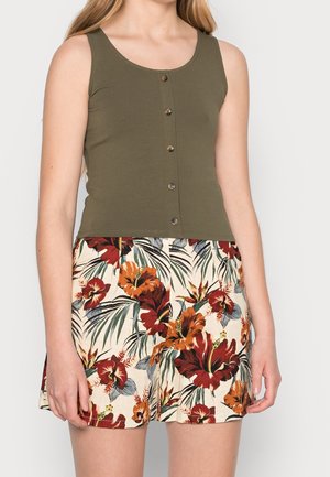 Kvinde iført en olivengrøn ærmeløs knapbluse kombineret med cremefarvede shorts med et rødt og orange tropisk blomsterprint.