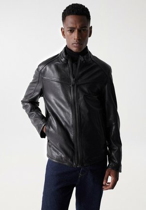 Chaqueta de cuero negra con cuello alto, cierre de cremallera y detalles de costura. Presenta mangas largas y un corte relajado, combinada con denim oscuro.