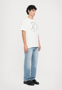 Ung mann står iført en hvit t-skjorte med grafikk, lyseblå jeans og svarte lærsko foran en ensfarget hvit bakgrunn.
