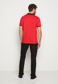 Rotes Poloshirt mit schwarzem Kragen und schwarzen Streifen an den Ärmeln, kombiniert mit schwarzen Hosen und schwarzen Sneakers. Der Hintergrund ist schlicht weiß.