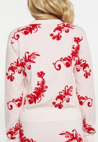 En blond person vänder bort och bär en ljusrosa tröja med djärva röda blommönster och swirl-mönster över ryggen och ärmarna.