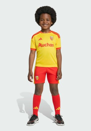Joven niño con afro lleva un uniforme de fútbol RCL amarillo y rojo, de pie y sonriendo frente a un fondo claro y liso.