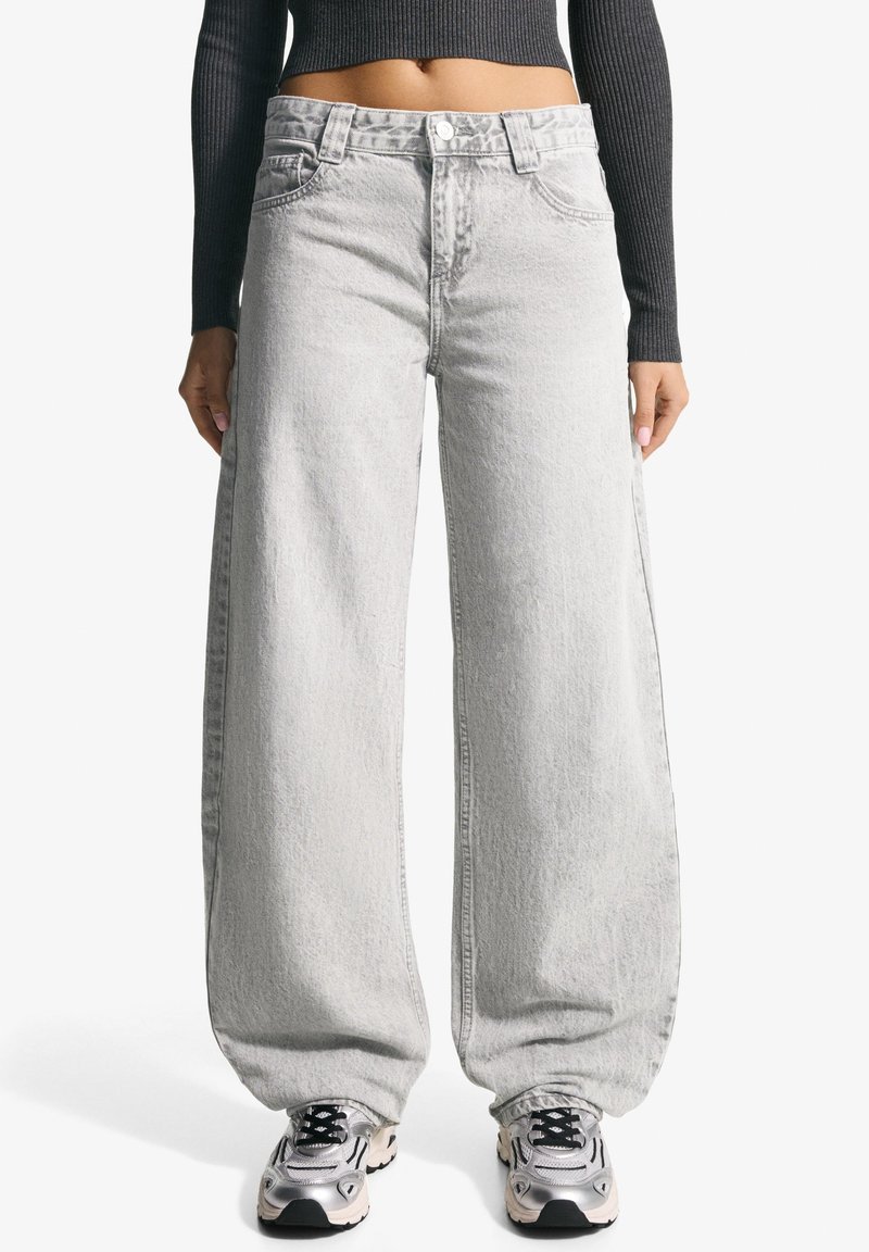 Femme portant un jean gris clair à jambes larges, un top noir court à manches longues et des baskets argentées, debout les mains détendues le long du corps.