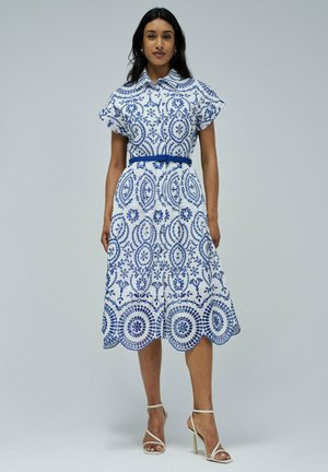 Robe blanche avec des motifs floraux bleus complexes, manches courtes, un col et une ceinture bleue assortie. Présente un ourlet festonné.