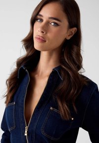 Granatowy kombinezon dżinsowy z zamkiem z przodu, spiczastym kołnierzem i dwoma kieszeniami na piersi. Złote okucia i widoczne przeszycia detalizują projekt.