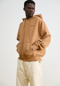 Brown hooded sweatshirt made from soft fabric, featuring a front pocket and ribbed cuffs. Paired with cream-colored trousers.