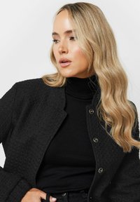 Svart texturerad jacka med krage, tryckknappar, bärs över en svart polotröja. Långt, vågigt hår synligt.