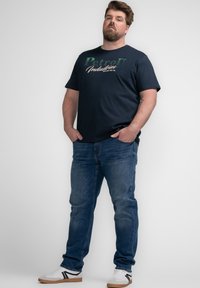 Camiseta de color azul marino oscuro con texto gráfico en verde y blanco "PetroD Industries." Combinada con jeans azules y zapatillas blancas con acentos negros.