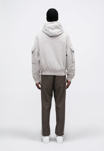 Persoană în picioare, cu spatele, purtând un hanorac gri deschis, pantaloni maro închis cu dungi pe laterale și adidași albi.