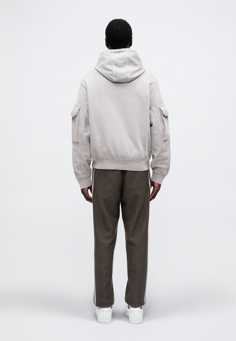 Person standing facing away wearing a light gray hooded sweatshirt, dark brown pants with side stripes, and white sneakers.