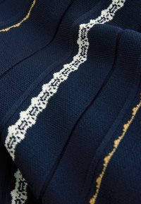 Tessuto a righe navy con bordo in pizzo bianco e accenti dorati, caratterizzato da un motivo ondulato che crea interesse visivo e profondità.