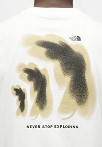 Πίσω μέρος λευκού πουκάμισου με τρεις μαύρες σιλουέτες χεριών που δείχνουν προς τα δεξιά, το λογότυπο "THE NORTH FACE" και το κείμενο "NEVER STOP EXPLORING" από κάτω.