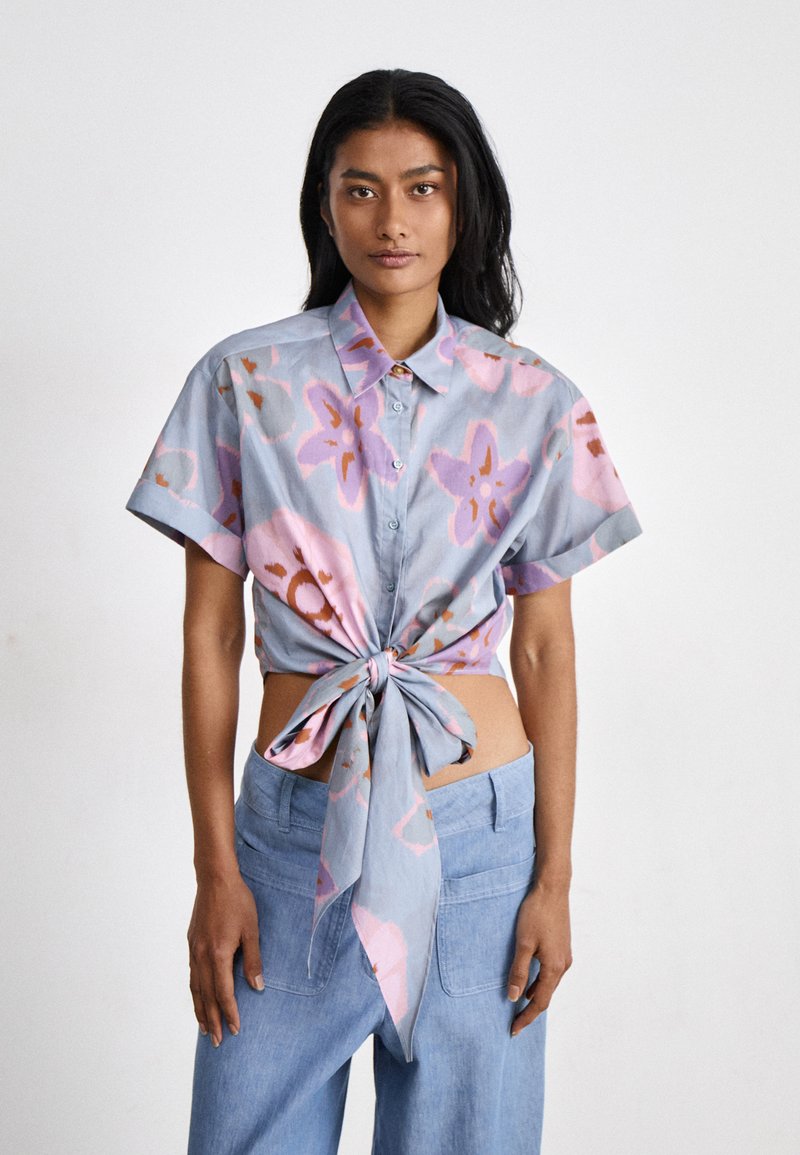 Camicetta corta a maniche corte di colore azzurro chiaro con pattern floreali in rosa e marrone. Legata in vita, con bottone sul davanti e colletto.