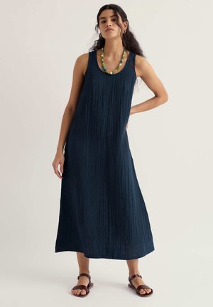 Vrouw met lang donker haar, gekleed in een mouwloze marineblauwe midi-jurk met textuur, een kleurrijke kralenhalsketting en bruine sandalen, staand met één hand op haar heup.