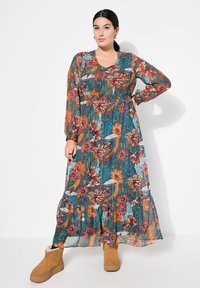 Maxikleid in Teal mit floralem Druck in Orange und Pink. Lange Ärmel, runder Ausschnitt, geraffte Taille und rüschenverzierter Saum. Stiefeletten in warmen Tönen.