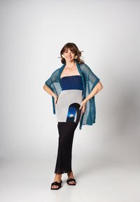 Turkusowa, przezroczysta dzianinowa kardigan na kolorowej sukience z blokami kolorów w odcieniach niebieskiego, szarego i czarnego, z okrągłym wzorem wycięć. Czarne sandały.