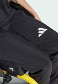 Pantaloni sportivi neri con una texture liscia presentano un logo geometrico bianco sulla coscia. La persona è seduta in una sedia a rotelle con un telaio giallo.