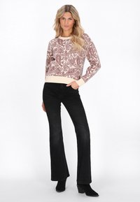 Donna bionda che indossa un maglione a paisley beige e marrone, jeans neri a zampa d'elefante e stivaletti neri, in piedi con una mano in tasca.