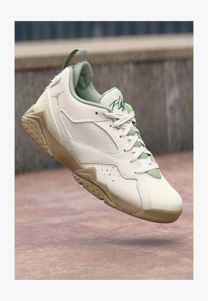 Zapatilla Air Jordan blanca y beige con paneles de malla, cordones y texto "Flight" en la lengüeta, suspendida sobre una superficie de hormigón rosa.