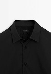 Black button-up shirt with a spread collar, featuring a smooth texture and two buttons on the front, labeled inside with brand and origin.