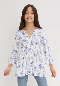 Tunika bianca floreale con motivi di fiori blu, scollo rotondo, chiusura con bottoni, maniche a campana e vita arricciata. Abbinata a jeans blu.