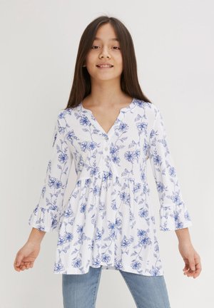 Tunika bianca floreale con motivi di fiori blu, scollo rotondo, chiusura con bottoni, maniche a campana e vita arricciata. Abbinata a jeans blu.