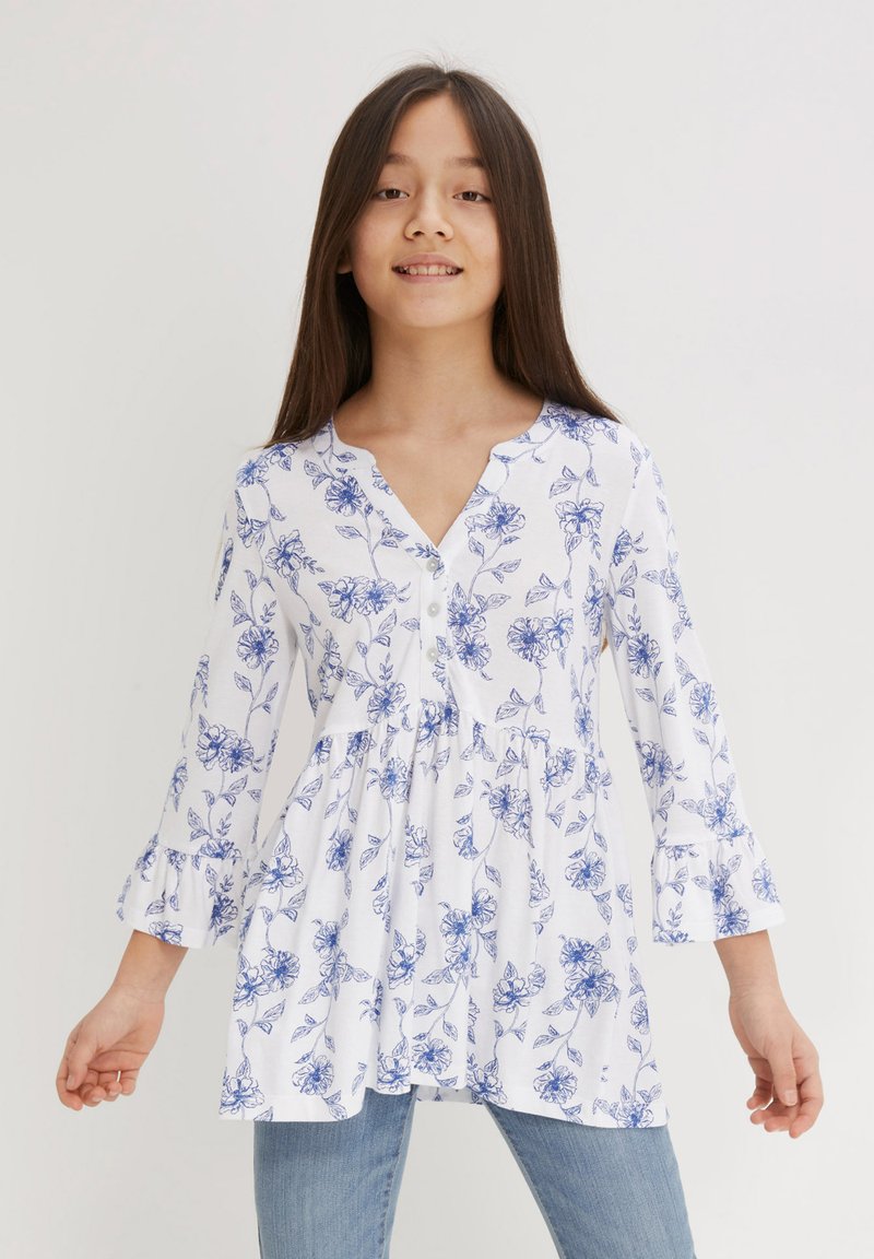 Tunika bianca floreale con motivi di fiori blu, scollo rotondo, chiusura con bottoni, maniche a campana e vita arricciata. Abbinata a jeans blu.