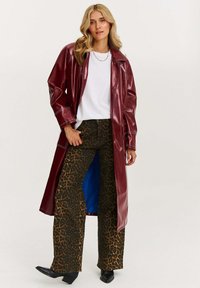Burgundská umelokožená dlhý kabát s kontrastným bielym stehom, kombinovaný s hnedými nohavicami s leopardím vzorom a čiernymi špicatými čižmami.